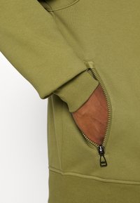 Main dans la poche d'une veste vert olive avec une fermeture éclair noire partiellement ouverte et un long poignet sur la manche.