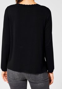 Svart långärmad topp med vågig halsringning, gjord av mjukt tyg. Baksidan har en slät textur och lös passform. Kombineras med grå jeans.