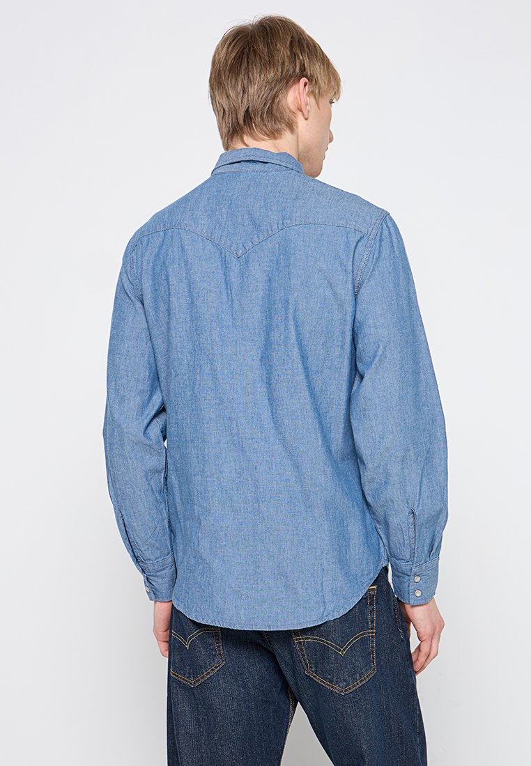 Camisa de mezclilla azul claro con mangas largas, dobladillo curvado y puños abotonados. Presenta un collar simple y costuras de hombro reforzadas.