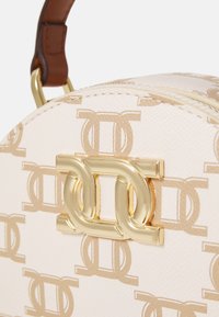 Kulatá krémová taška s texturovaným povrchem, kterou zdobí zlaté logo dvojitého C a opakující se světle béžový vzor. Hnědá kožená rukojeť.