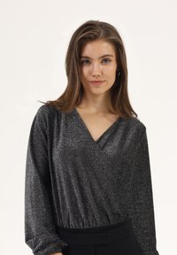 Svart glitrende omslagsbluse med lange ermer, dyp V-hals og rynket midje, kombinert med svarte bukser. Glatt tekstur og skinnende detaljer.