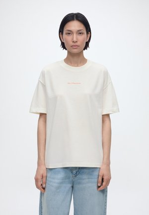 Persona con capelli corti e scuri indossa una maglietta bianca larga con un piccolo testo arancione e jeans azzurri, in piedi davanti a uno sfondo bianco uniforme.