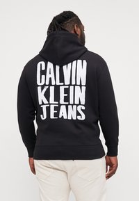 Black hoodie with a large white "CALVIN KLEIN JEANS" logo on the back. Features a drawstring hood and ribbed cuffs and hem.