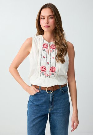 Femme debout portant un chemisier blanc sans manches avec une broderie florale rouge, un jean bleu et une ceinture tressée marron sur un fond uni.