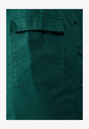 Tamnozelena tkanina košulja s džepom na prsima, glatke teksture, zatvaranjem na gumbe i suptilnim sjajem. Šivani detalji duž rubova.