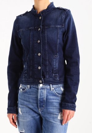 Kobieta ubrana w granatową, zapinaną na guziki kurtkę dżinsową z pagonami oraz jasnoniebieskie, postrzępione dżinsy na białym tle.