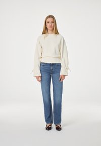 Vrouw met lang blond haar die een crèmekleurige sweatshirt draagt met gestrikte manchetten, blauwe rechte jeans en donkere schoenen met spitse neuzen, staat recht vooruit.