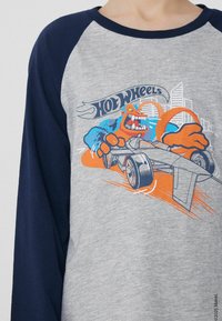 Šedá košile s dlouhým rukávem a námořnickými raglánovými rukávy, s barevným karikováním grafiky monstra a auta s textem "Hot Wheels" nahoře.
