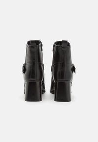 Botas de cuero negro hasta el tobillo con un alto tacón cuadrado, cierre de cremallera lateral y detalle decorativo de hebilla en el lado exterior. Textura suave.