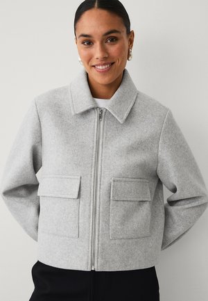 Woman smiling, wearing a light gray wool zip-up jacket with large flap pockets and a wide collar, standing against a plain background.