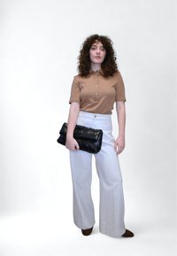 Καφέ κοντομάνικο πουκάμισο με κουμπιά, λευκό παντελόνι με φαρδιά πόδια και μαύρος πλεκτός φάκελος. Καφέ παπούτσια, σγουρά μαλλιά, ελάχιστο φόντο.