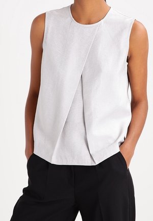 Mouwloos lichtgrijs bovenstuk met een asymmetrisch voorpaneel, gedragen met een zwarte broek, handen in de zakken, tegen een effen achtergrond.