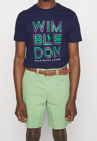 Marineblå bomullströja med "WIMBLEDON"-grafik i grönt och lila, tillsammans med mintgröna shorts och ett brunt vävt bälte.