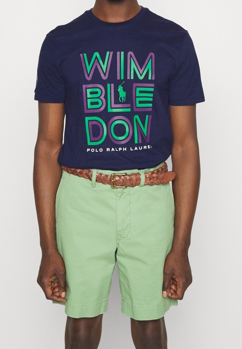 Marineblå bomullströja med "WIMBLEDON"-grafik i grönt och lila, tillsammans med mintgröna shorts och ett brunt vävt bälte.