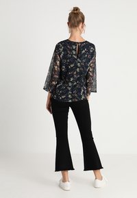 Blusa floreale con manicheTrasparenti a campana, sfondo blu navy e dettaglio a chiave sul retro, abbinata a pantaloni neri a campana e scarpe bianche.
