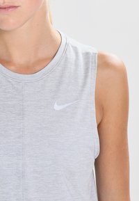 Camiseta deportiva sin mangas de color gris claro con escote redondo y un logo blanco de Nike en el pecho izquierdo. Textura de tela suave y diseño casual.