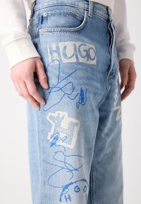 Light blue denim jeans featuring blue and white graffiti-style text and designs, with a relaxed fit and prominent stitching details.