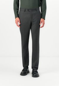 Man wearing dark gray pinstripe trousers, black loafers, and a long-sleeve green shirt, standing against a white background.