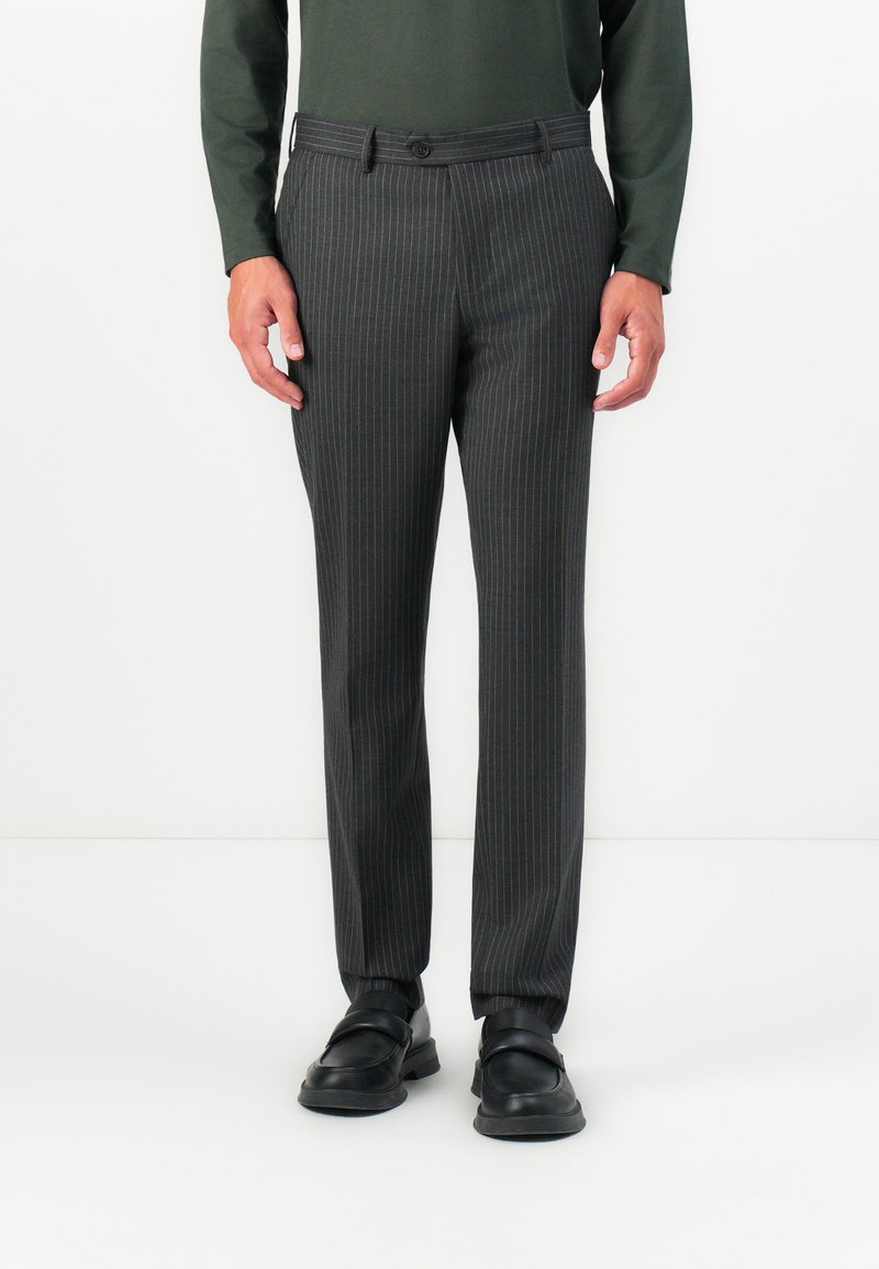 Man wearing dark gray pinstripe trousers, black loafers, and a long-sleeve green shirt, standing against a white background.