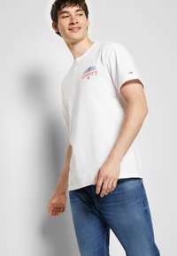 Hvid bomulds T-shirt med et trykt rødt og blåt design med teksten "TOMMY'S" og et logo. Korte ærmer og rund hals.