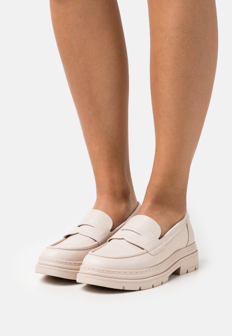 Mocassins en cuir beige clair avec semelles épaisses portés pieds nus, montrés de face et de côté sur un fond uni clair.