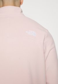 Pullover in felpa rosa chiaro con colletto alto e logo bianco ricamato sul retro. Presenta cuciture lungo la parte superiore della schiena e sulle spalle.