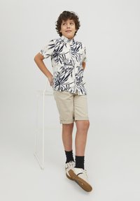 Camicia bianca a maniche corte con motivo di foglie blu navy, pantaloni corti beige arrotolati, calze nere e sneakers bianche con suole marroni. Sfondo chiaro.