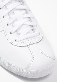 Vit sneakers med slät läderöverdel, platta vita snören, texturerad gummisula och synliga sömmar längs tån och sidorna.