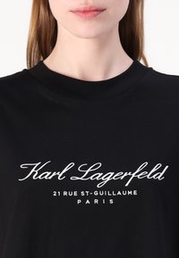 Juodas marškinėlis su apykakle tipo crew. Su baltais siuvinėtų žodžių įrašais: "Karl Lagerfeld 21 RUE ST-GUILLAUME PARIS." Lygus audinys.