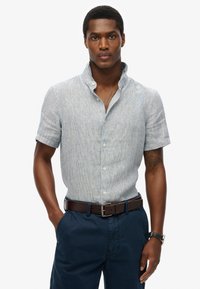 Camisa de manga corta, abotonada, en azul claro con un sutil patrón de rayas. Cuenta con un collar tipo stand y una tela suave. Combinada con pantalones oscuros y un cinturón.