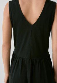 Schwarzes ärmelloses Kleid mit tiefem V-Ausschnitt, weichem Stoff, gerafftem Taillenbereich und schwingendem Rock, das ein einfaches und minimalistisches Design präsentiert.