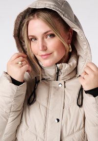 Beige pufferjack met een gewatteerd ontwerp, afneembare capuchon en contrasterende zwarte binnenkant. Heeft metalen drukknoopsluitingen en trekkoordaccenten.