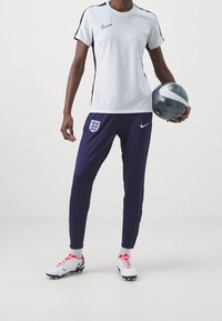 Camiseta blanca de manga corta de Nike con detalles negros, pantalones púrpura de Nike con un escudo de Inglaterra, y botas blancas con detalles rojos, sosteniendo un balón de fútbol gris.
