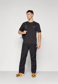 Jauns vīrietis melnā T-kreklā un biksēs, ar melnu mugursomu pār vienu plecu, stāv pret vienkāršu gaišu fonu.