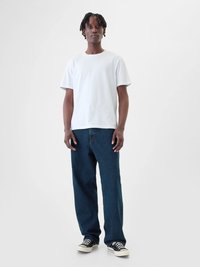 Camiseta de algodón blanca de mangas cortas, combinada con jeans de mezclilla azul oscuro. Zapatillas deportivas con diseño en blanco y negro, de pie contra un fondo neutro.