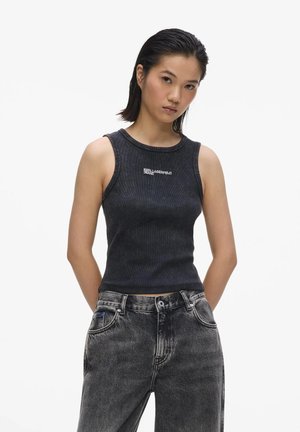 Mladá žena oblečená v čiernom tielku s nápisom „Karl Lagerfeld Jeans“ a vybledlých čiernych džínsoch, stojaca s rukami za chrbtom.