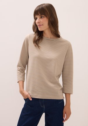 Femme aux cheveux châtains moyens portant un haut à rayures beige et blanches à manches longues et un pantalon bleu marine, main dans la poche, regardant sur le côté.