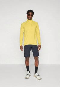 Gul topp med långa ärmar och halv dragkedja; kombinerad med mörka shorts. Modellen bär solglasögon och lätta vandringsskor. Mörka strumpor kontrasterar mot skorna.