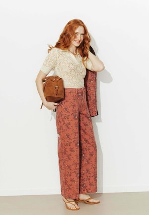 Femme aux cheveux roux souriante, portant un haut en dentelle crème, un pantalon à jambes larges à fleurs rouille, tenant un sac en daim marron et une veste sur l'épaule, en sandales.