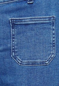 Denimtyg i medelblå färg med en rektangulär bakficka med sömdekor och en subtil textur.