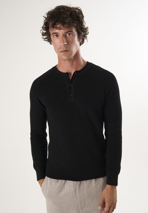 Schwarzes gestricktes Langarm-Henley-Shirt mit einem geknöpften Ausschnitt und geripptem Muster, kombiniert mit heller Hose.