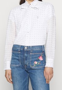 Blouse blanche avec des motifs bleus, col noué et manches longues, associée à un jean taille haute bleu avec une broderie florale sur la poche.