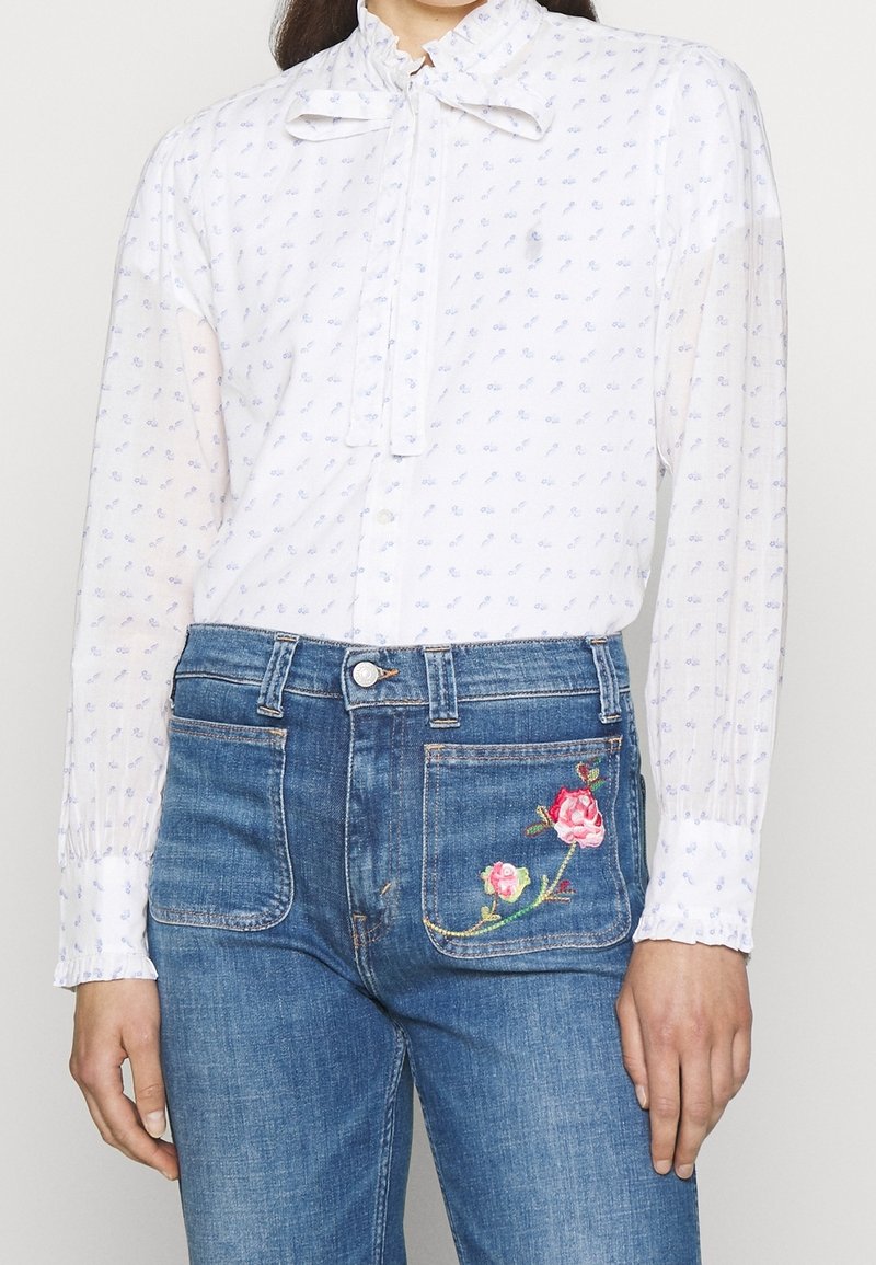 Blouse blanche avec des motifs bleus, col noué et manches longues, associée à un jean taille haute bleu avec une broderie florale sur la poche.
