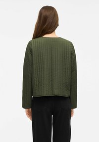 Vrouw met lang bruin haar draagt een gewatteerde olijfgroene jas en zwarte broek, weergegeven vanachter tegen een witte achtergrond.