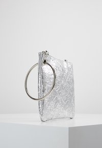 Silverfärgad, krinklad metallhandväska med en stor silverringhandtag. Har en rektangulär form och en slät yta. Dragkedjestängning på toppen.