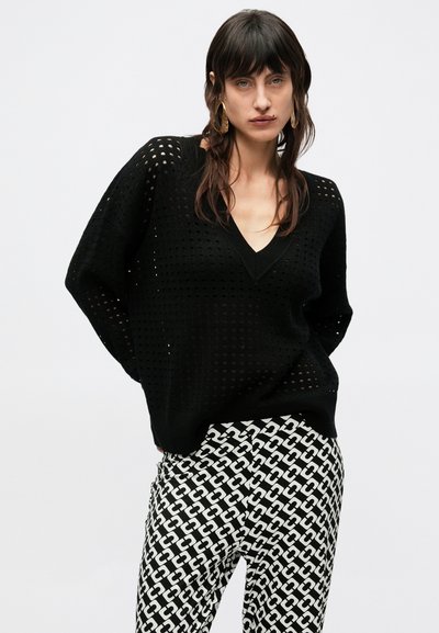 Čierny pletený sveter s výstrihom do V a vzorom otvoreného pletenia, doplnený bielymi a čiernymi nohavicami s geometrickým vzorom spojení.