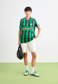 Žalia ir juoda juostelėta maikutė su ryškiu grafiniu logotipu, derinama su baltais smulkiais juostelėtais šortais ir žaliais akcentais ant batų bei kojinių.