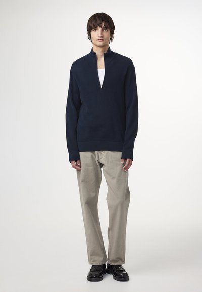 Navy pullover met een kwart rits, ribbelstructuur, contrasterende manchetten, gecombineerd met lichtbeige broek en zwarte schoenen.
