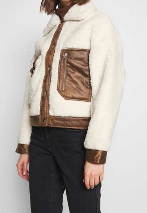 Witte sherpa-jas met bruine leren rand, een borstzak met rits en drukknoopsluiting, gedragen met een zwarte spijkerbroek.