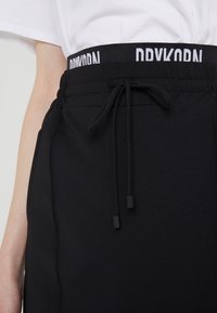 Pantalons de jogging noirs avec une taille élastique, ornés d'une marque blanche contrastante, de cordons de serrage avec des embouts noirs et d'une texture de tissu lisse.